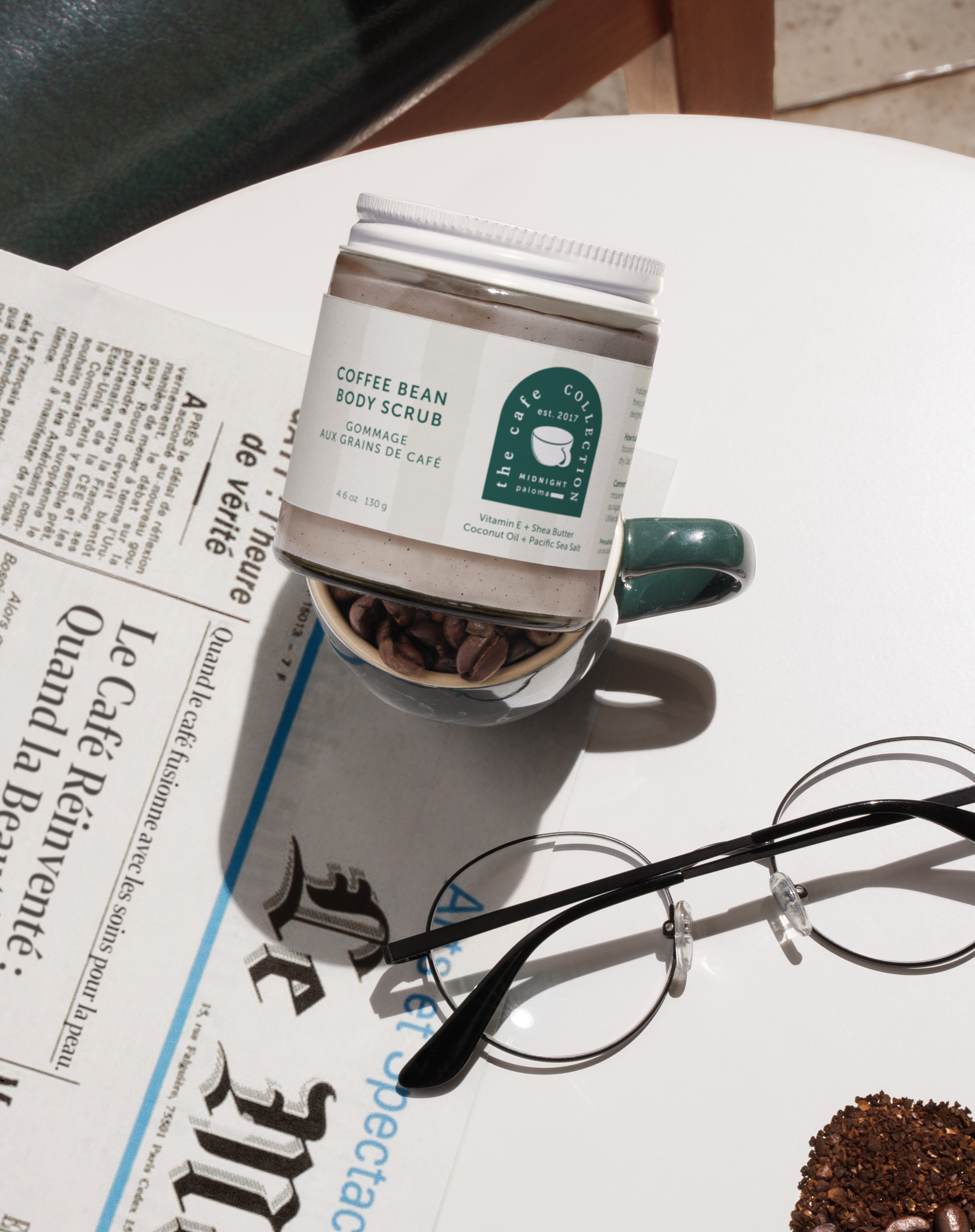 Danielle Gracioso Graphic Designer Vancouver BC Canada – Styled lifestyle photo of Coffee Bean Body Scrub resting in a coffee cup on a table, beside a French newspaper and eyeglasses. Shot and styled by Danielle Gracioso.